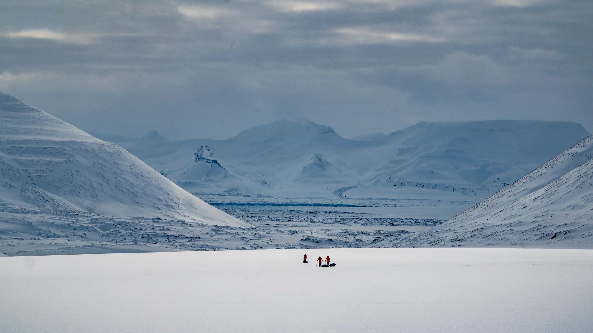 Snow-covered mountains frame a vast white plain with three small figures traveling across, likely on sleds or skis.